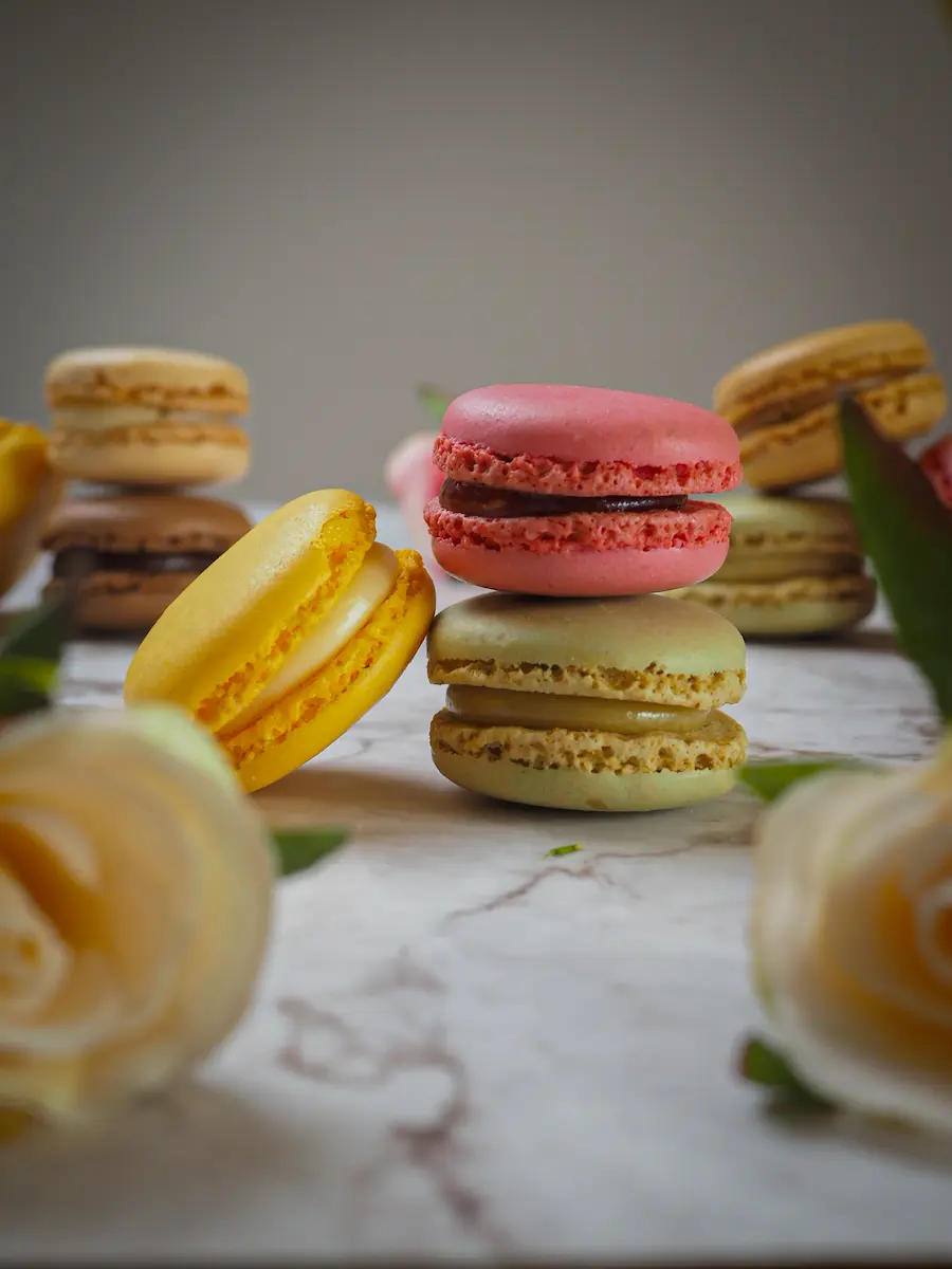 Assorted gourmet French macarons in pink, yellow, and green flavors, elegantly plated with floral accents on a marble background