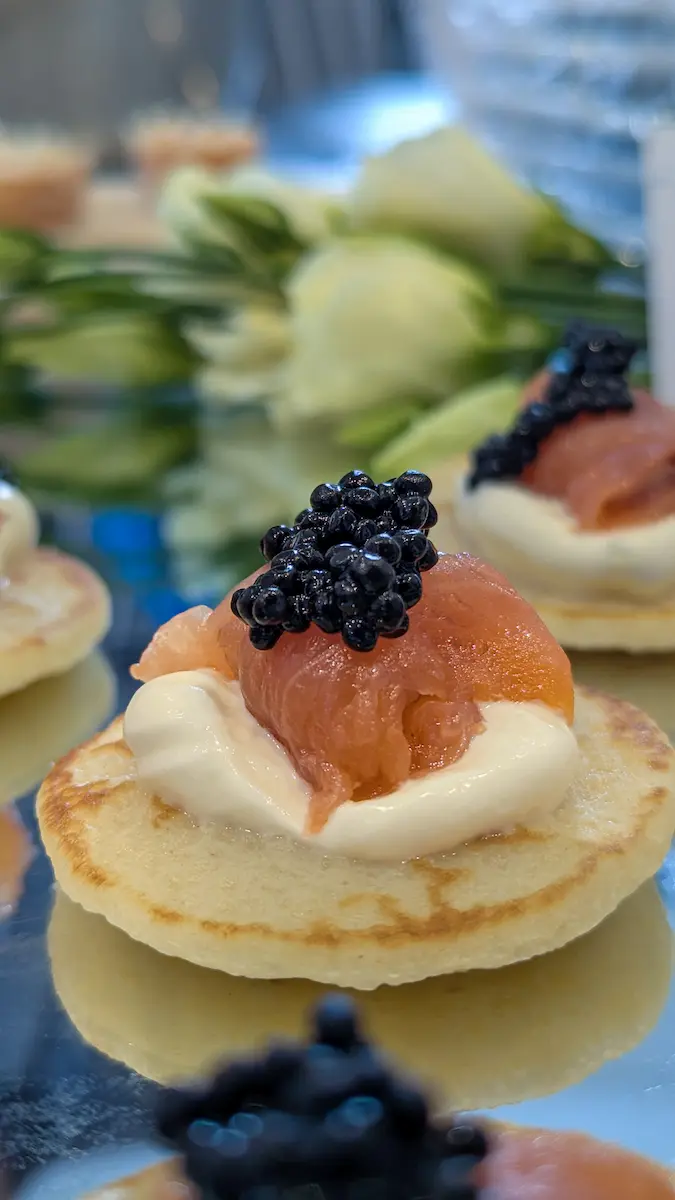 A close-up photograph captures a blini canapé topped with a dollop of white creme fraiche, a folded slice of smoked salmon, and a generous mound of black caviar