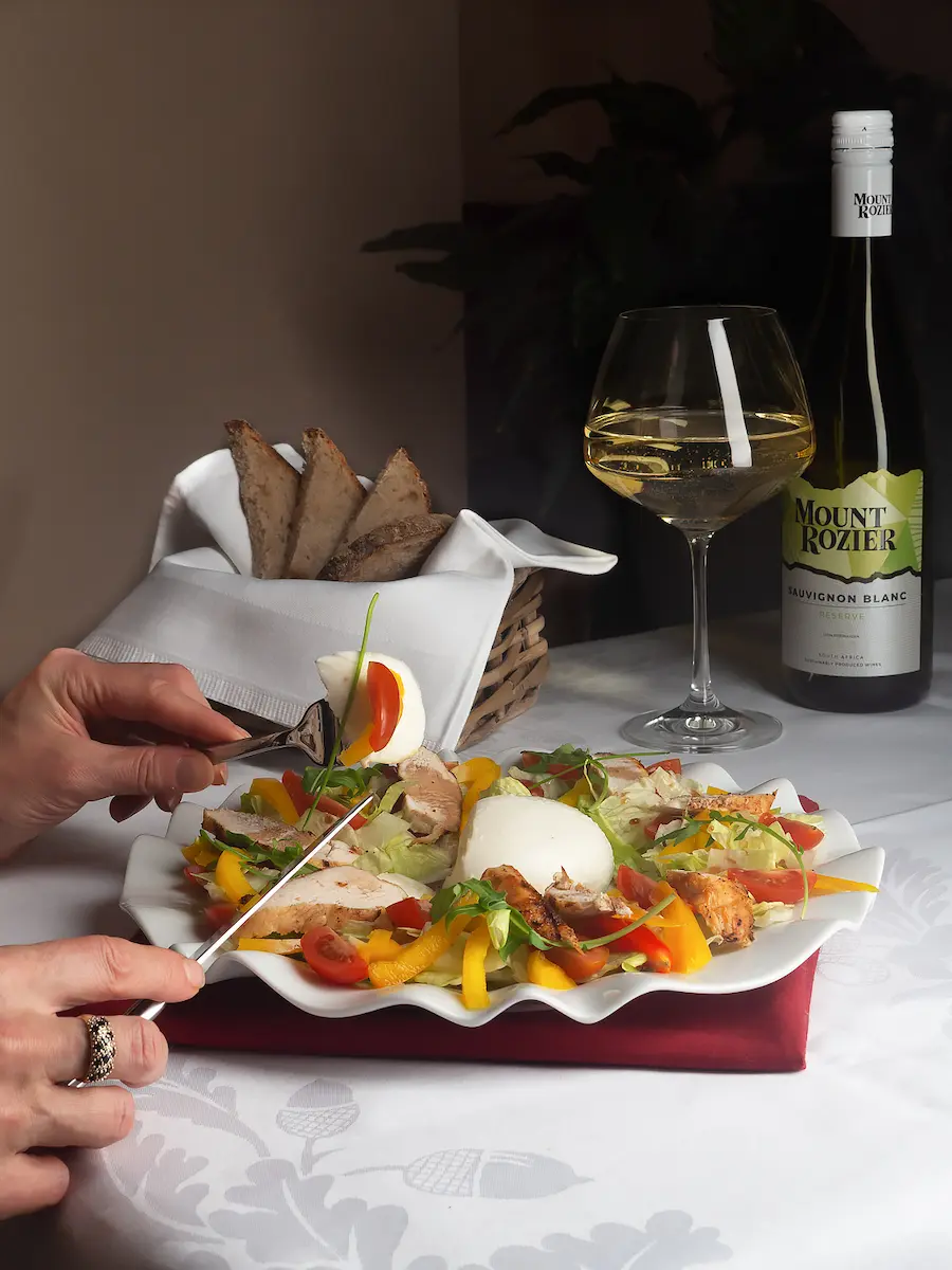 A diner's hands use a fork and knife to eat a colorful salad featuring grilled chicken, yellow and red bell peppers, cherry tomatoes, and a large ball of fresh mozzarella or burrata on a white wavy plate