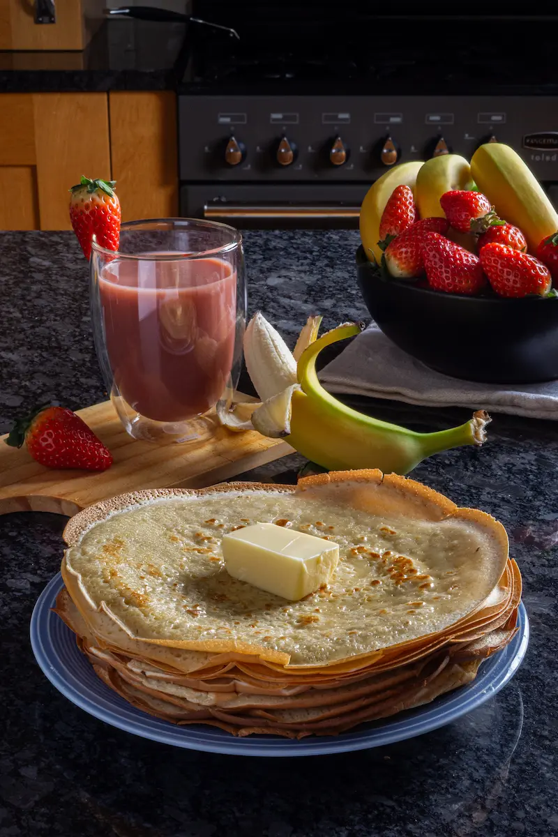 Stack of thin crepes topped with butter, served with a fresh strawberry smoothie and fruit bowl