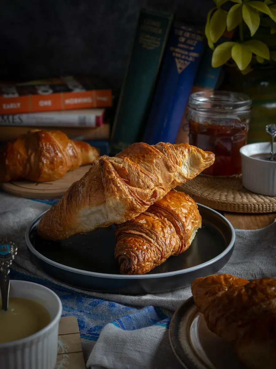 Golden flaky croissants plated next to vintage books and a jar of jam for a cozy breakfast theme