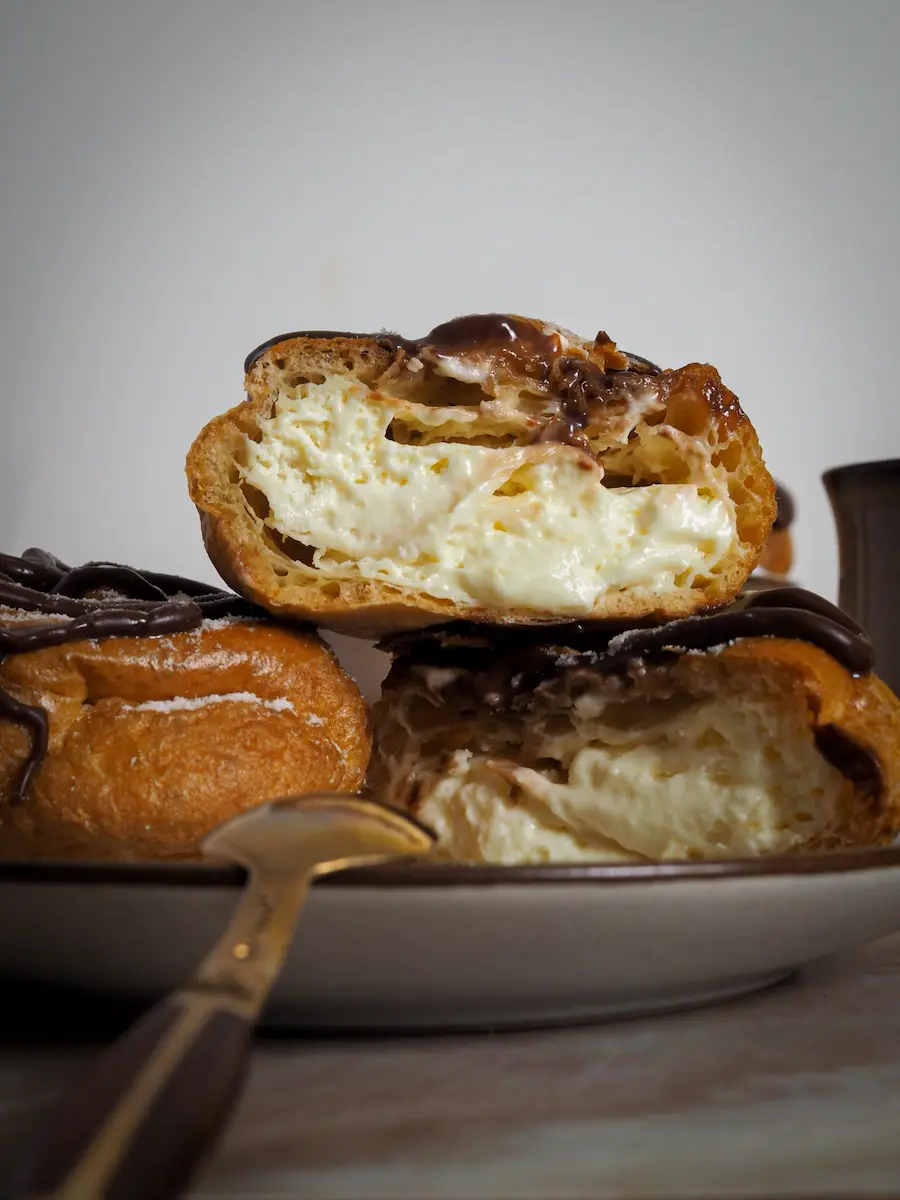 Cut choux pastry filled with whipped cream and topped with chocolate ganache, highlighting the airy interior texture of the profiterole
