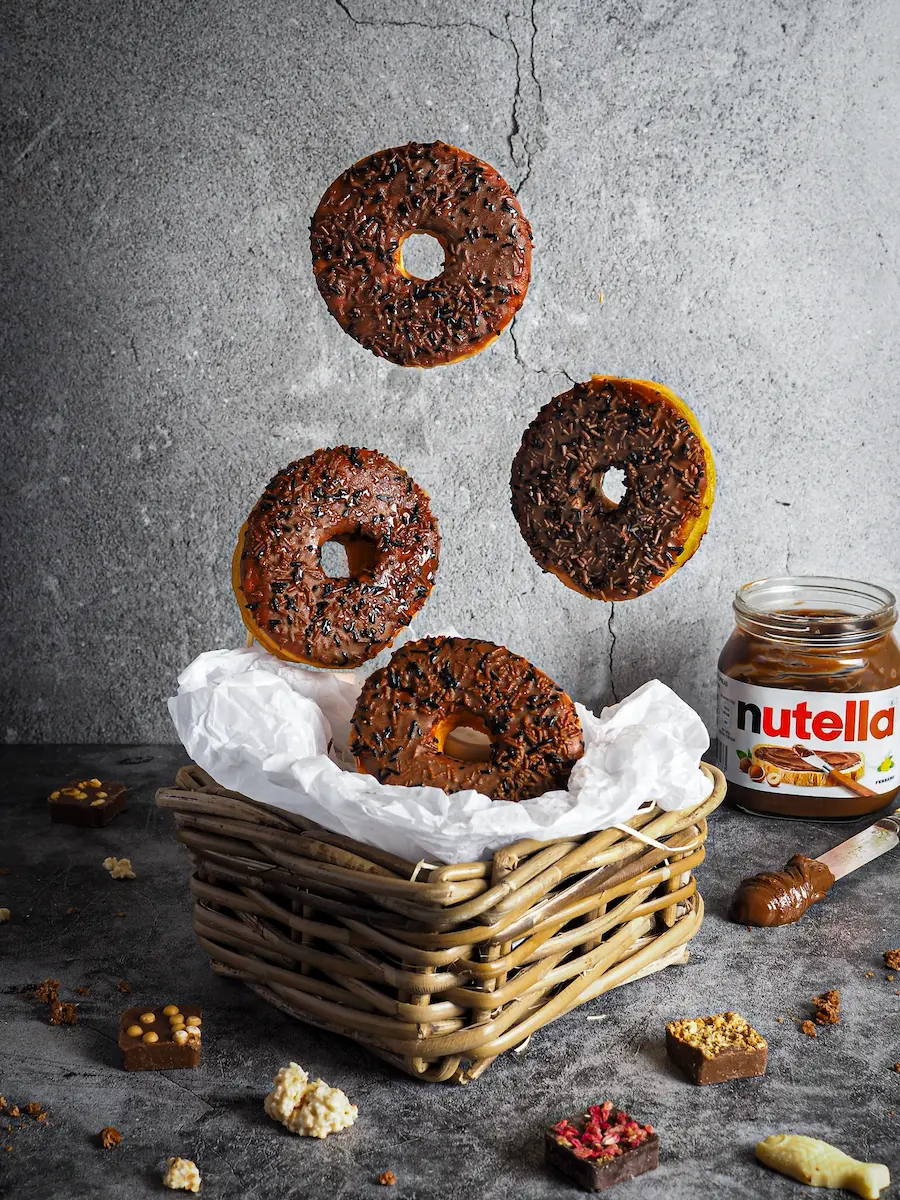 Creative food photography of floating chocolate sprinkle donuts hovering over a basket near a Nutella jar