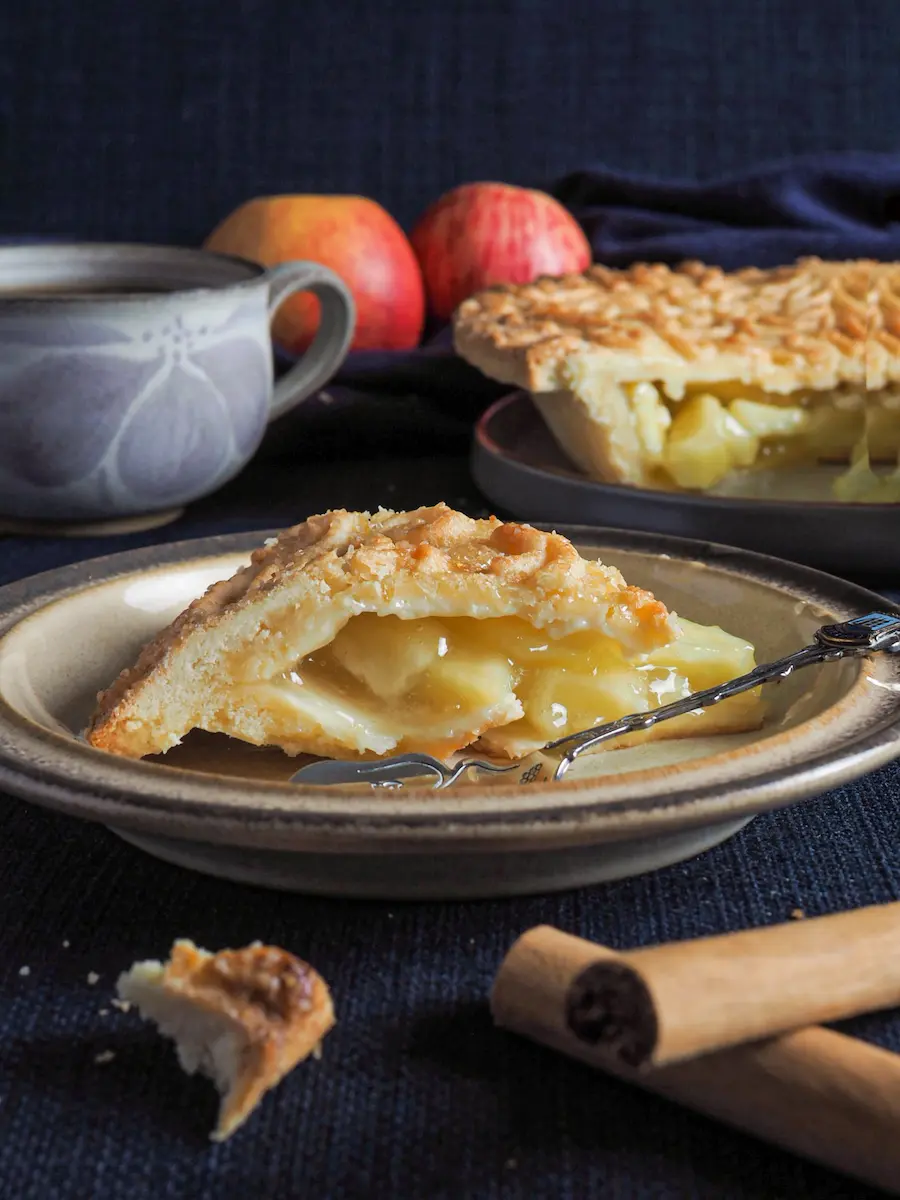 Rustic apple pie slice with lattice crust and gooey filling, plated with cinnamon sticks and a cup of tea in a cozy setting