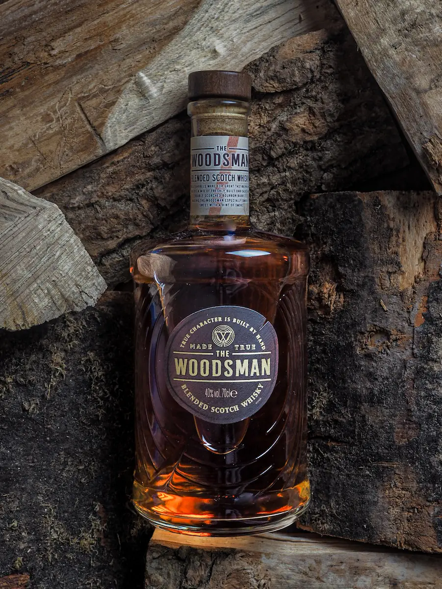A bottle of The Woodsman Blended Scotch Whisky nestled among stacked rough-hewn wooden logs